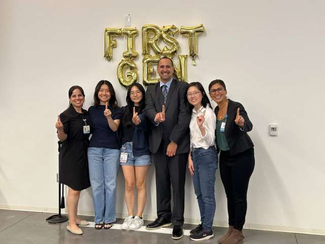 Seven people smiling with thumbs up beneath gold 'FIRST GEN' balloons