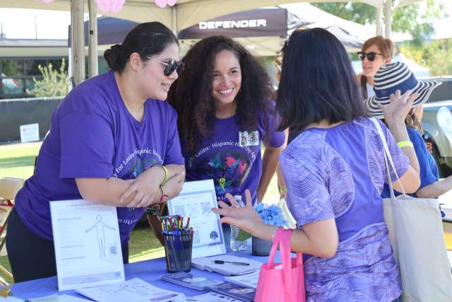 Four people at an outdoor information table under a tent, discussing pamphlets