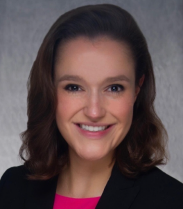 Professional headshot of a person with short wavy hair, wearing a dark blazer over a bright pink top, facing forward against a gray studio background.