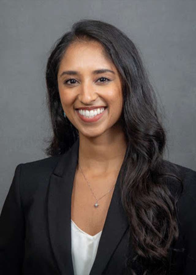 Professional headshot: smiling woman in black blazer on gray background.