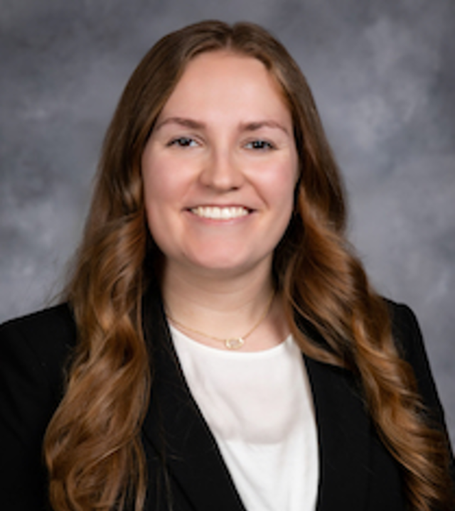 Professional headshot of a person with long wavy hair, wearing a black blazer over a light-colored top and a small necklace, facing forward against a gray studio background.