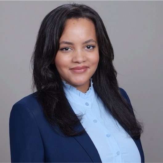 A smiling woman with dark hair and a blue shirt in front of a gray background.