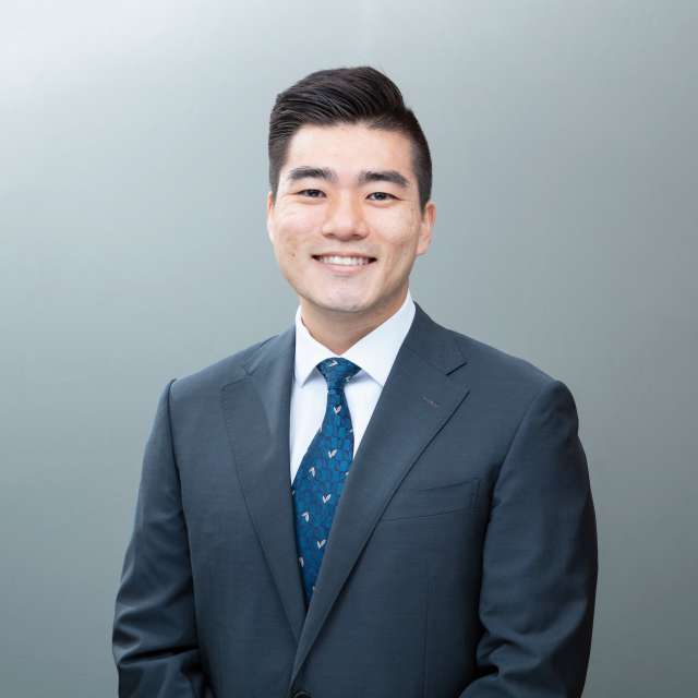  A headshot of Andrew Pak, a smiling man with dark hair, wearing a dark suit and a blue patterned tie against a grey background.