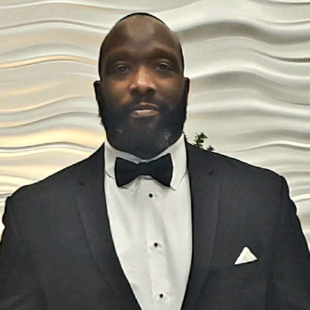 Portrait of Courtney Shepherd in a tuxedo and bow tie, standing in front of a modern wavy-textured wall at a formal UCLA Health event.