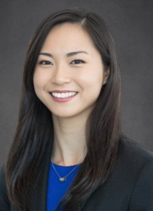 Professional headshot of a person with straight shoulder-length hair, wearing a dark blazer over a bright blue top and a necklace, facing forward against a dark neutral background.