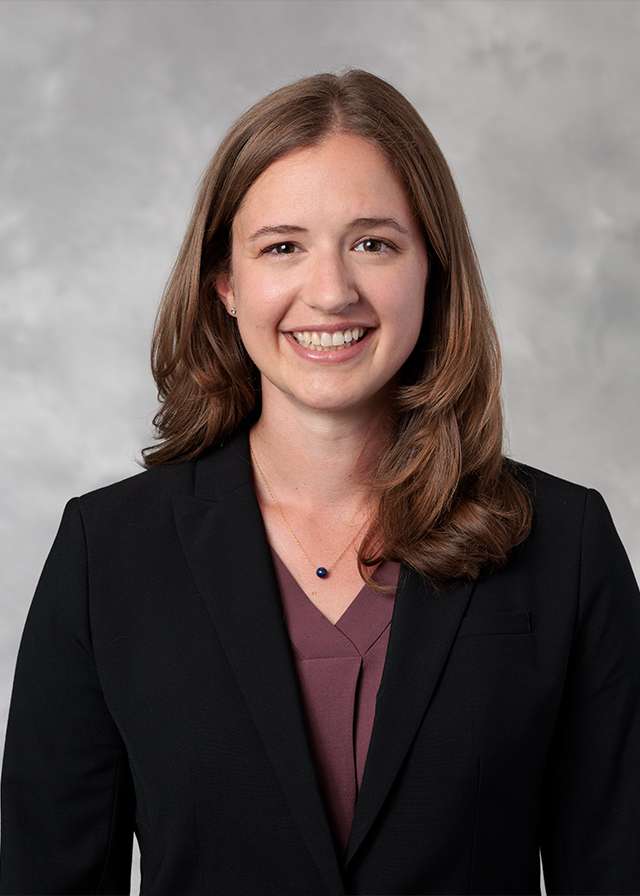 Smiling woman in black blazer and purple top against soft gray background