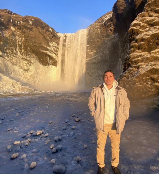 Person standing in front of a large waterfall surrounded by rocky terrain.
