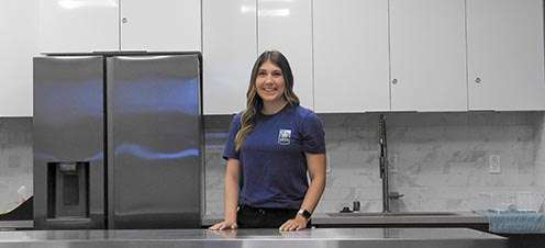 Woman standing in a modern kitchen beside a stainless steel refrigerator and dark countertop.