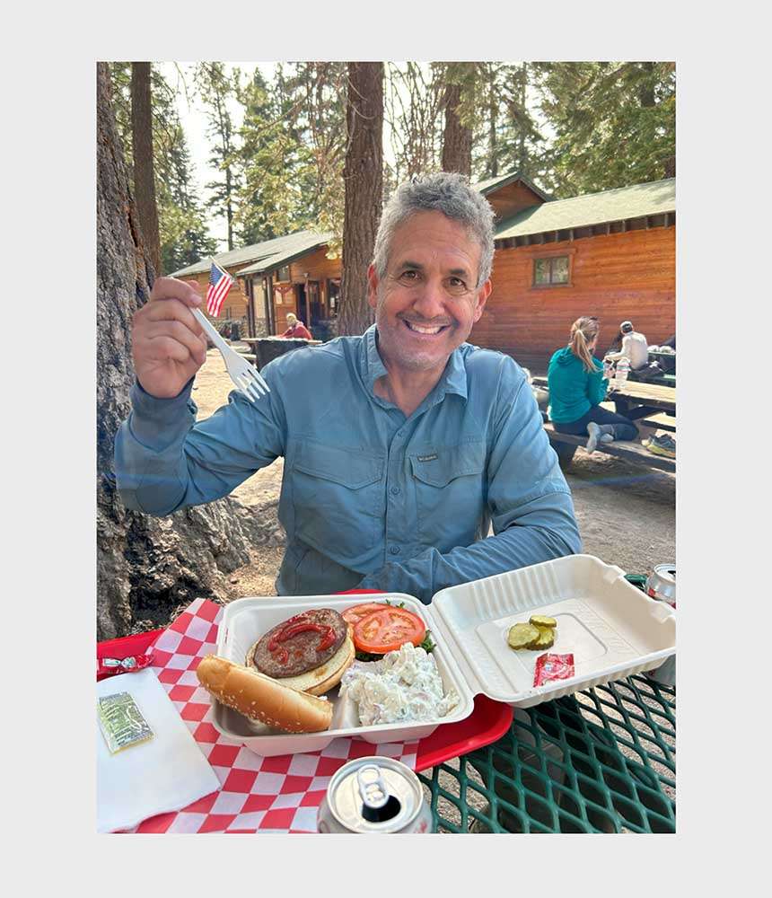 Michael Teitell enjoying a picnic meal with a burger, sides, and a scenic background.