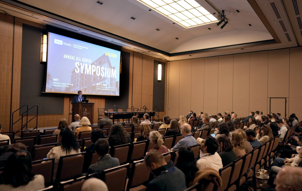 Dr. Michael Teitell speaking at the UCLA Health Jonsson Comprehensive Cancer Center Symposium 2024, standing at a podium with a presentation screen behind him.