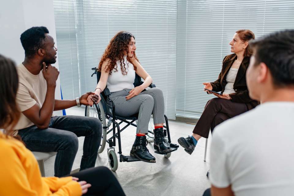 A group of people sit in a circle talking, including a woman in a wheelchair, in a bright indoor setting.