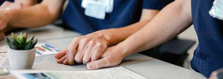 Two hands clasped together on a table, with a small potted plant nearby.