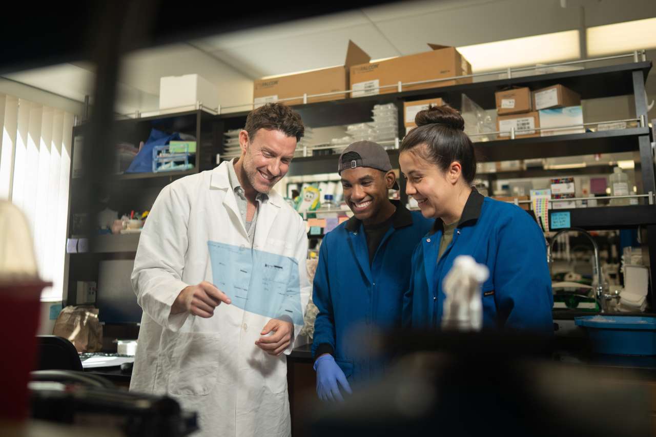 Researcher in lab coat reviewing clipboard with two colleagues in laboratory
