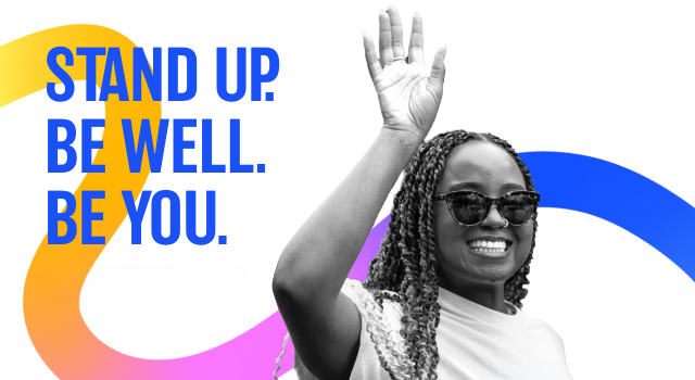 A woman in black and white waving in front of a stripe of color, in gradient from yellow to blue, with the text 'Stand up. Be well. Be you.'