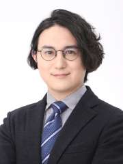 A headshot of Koichiro Kimura with dark, wavy hair, round glasses, and smiling, wearing a dark suit, light shirt, and striped tie.