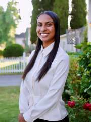 A professional headshot of Lela Theus, a smiling woman with long, straight dark hair, wearing a white long-sleeved shirt outdoors.