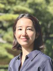 Woman with shoulder-length hair wearing a dark striped shirt, outdoors in a park.