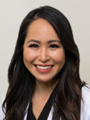 Joanna Wong with long dark hair wearing a white coat and black top, smiling against a plain background.