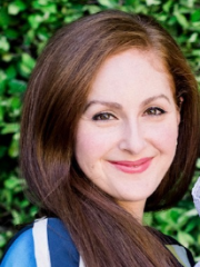 Nicole Shoraka with long brown hair wearing a blue top, smiling in front of greenery.