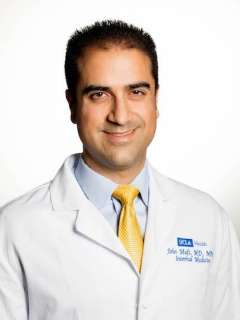 A professional headshot of Dr. John Mafi, a man with dark hair, smiling in a white lab coat with a gold tie against a light background.