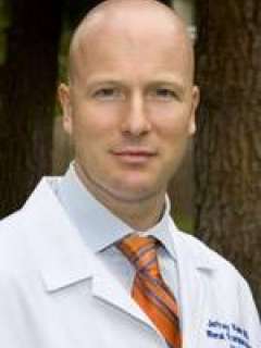 Jeffrey in a white lab coat with an orange striped tie, outdoors among trees.