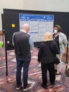 Three people stand looking at a large scientific poster displayed on a black board during a conference or meeting.