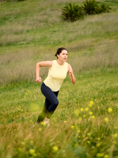 woman with brown hair and a yellow tank top, running in a grass field