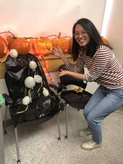  An-Chieh Feng smiles and points to a chair decorated with a black trash bag and white spherical lights. Carved pumpkins sit on a table behind her.