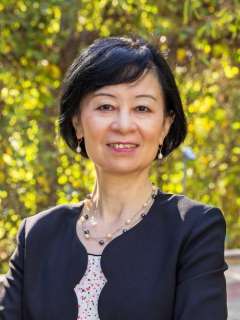 Person wearing a dark blazer over a patterned blouse with a pearl necklace, standing outdoors in front of greenery.