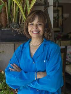 Gail Wyatt a bright blue shirt stands indoors with arms crossed, smiling in front of potted plants.
