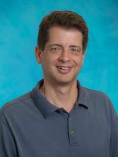  A portrait of Dr. Stephen Smale, with short brown hair, smiling in a blue polo shirt against a blue background.