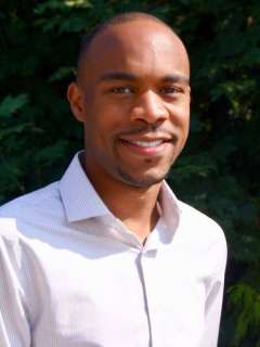 Julian McNeil in a white shirt stands outdoors against a green background.