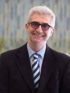 Dr. Emanuel Maidenberg in a suit with a striped tie, blurred face, standing against a soft background.