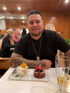 Person at a restaurant table with a chocolate cake topped with berries and a lit candle beside a flower-garnished dessert, with drinks nearby on table
