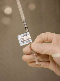 Gloved hand holds a syringe labeled “COVID-19 Vaccine” with dosage and expiration date visible.
