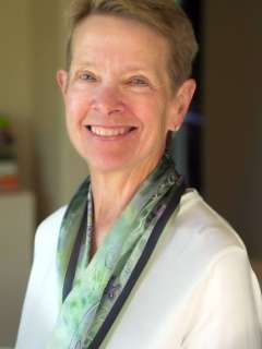Headshot of Laurie Adami, a woman with short, light hair and a scarf, smiling gently at the camera. Her pose implies she is kind and approachable.
