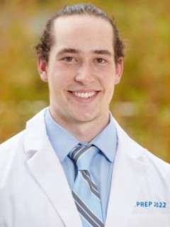 Nathan Ballew with brown hair tied back, wearing a white coat, a blue shirt, and a striped tie.