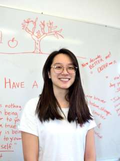  A smiling An-Chieh Feng with long dark hair and glasses stands in front of a whiteboard with handwritten notes and a tree drawing.