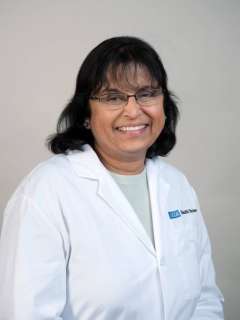 A smiling woman, Dr. Sherin U. Devaskar, with dark hair, wears a white medical lab coat. Her warm expression suggests kindness and expertise.