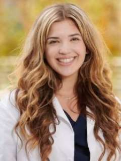 Alexxandra Hurtado with long wavy hair wearing a white coat smiles outdoors with trees in the background.