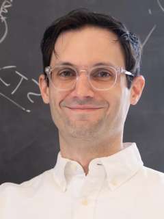 Dr. Gregory Donaldson stands with glasses and white shirt smiling in front of a chalkboard with equations
