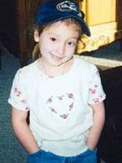 Child wearing a blue baseball cap, a white shirt with floral heart design, and jeans with hands in pockets.
