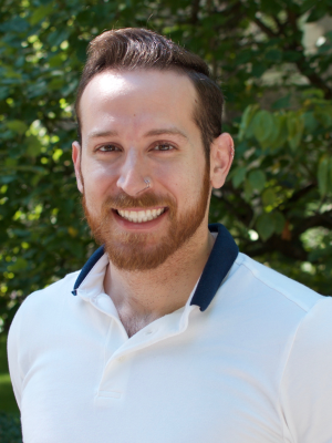 Smiling bearded man in white polo shirt outdoors with green foliage