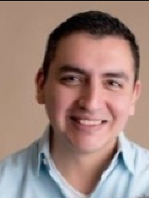 A man with short brown hair, wearing a blue collared shirt, smiling in front of a beige background