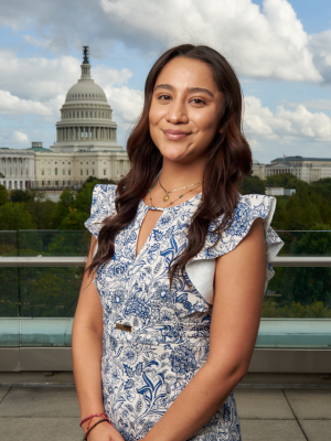 A woman with long wavy brown hair, wearing a floral dress, posing in front of the US Senate