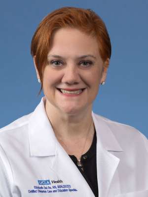  Red-haired woman, Elizabeth Del Re, smiling and wearing a white lab coat, in a friendly and welcoming headshot against a blue background.