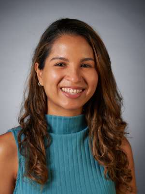 A woman with long brown wavy hair, wearing a teal sweater, smiling in front of a blank background