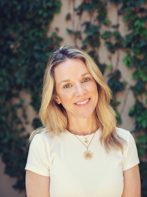Blonde woman in a white shirt standing in front of green foliage.
