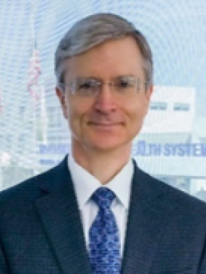 A man with short grey hair, wearing glasses with a suit and tie, posing in front of a building window
