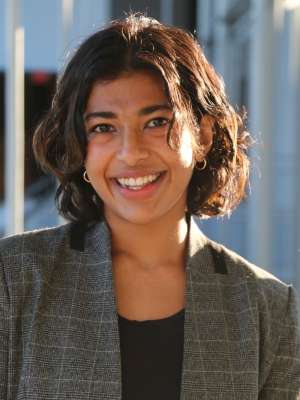 Smirthy Ganesan with short, curly hair wearing a gray blazer, standing indoors.
